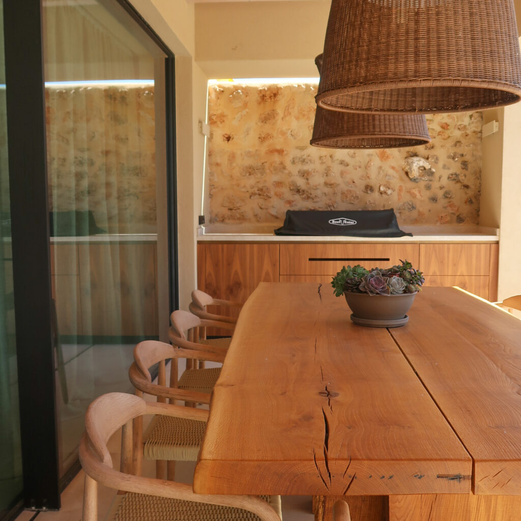 Solid wood exterior dining table. Kave Home chairs in beige cord. Bespoke barbecue unit with a Beefeater ProLine gas bbq, Bover pendant lights above the dining area.