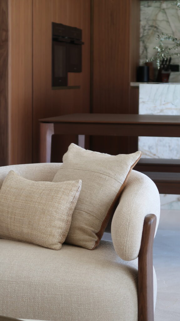 Kave Home armchair in beige upholstery and walnut carpentry. A dining table from Ethnicraft behind it, and a bespoke kitchen beyond in the background.